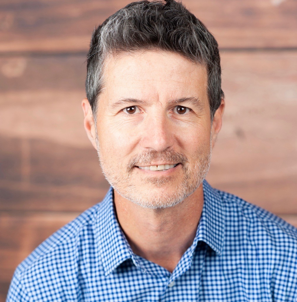Dave DeFazio, Chief Strategy Officer at StrategyCorps, smiling in a blue checked shirt against a warm wood background